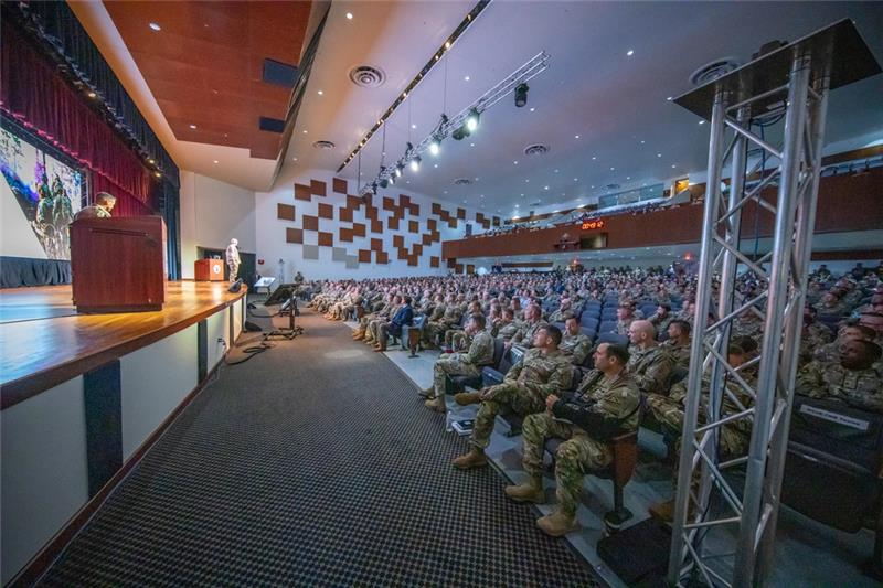 A conference room, showing a speaker on a stage and military members in the audience