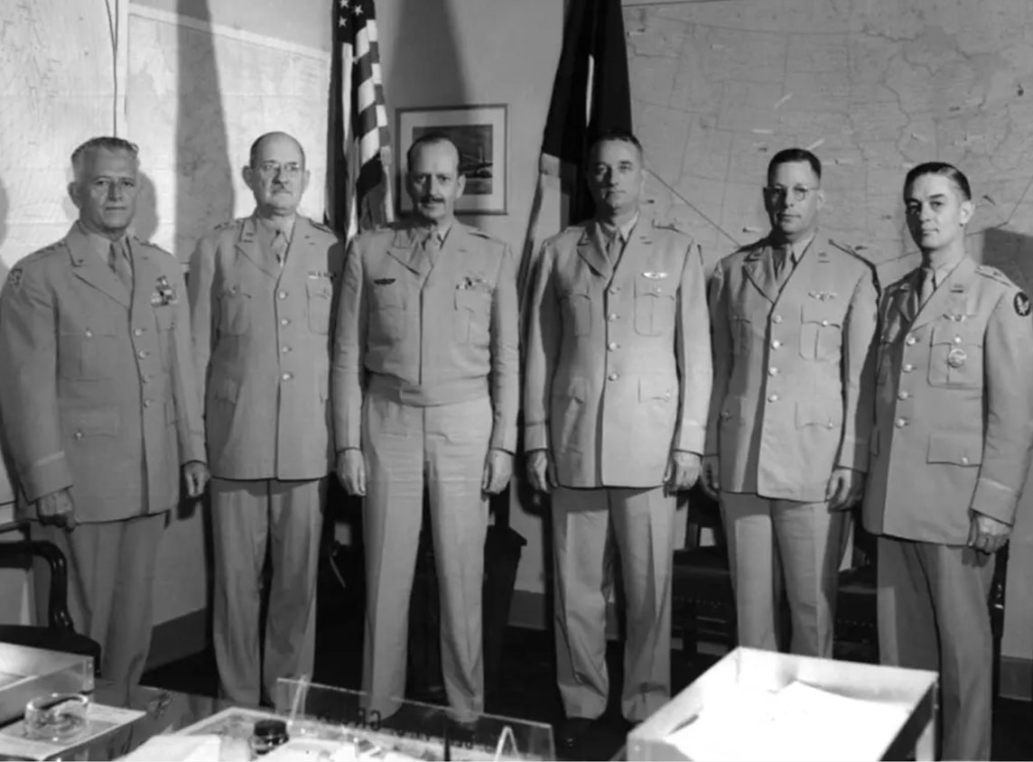 Six men in military uniforms standing in front of American flat