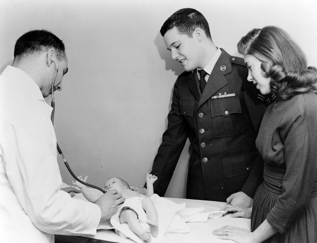 A soldier and his wife look down on their baby, who's being examined by a doctor with a stethoscope