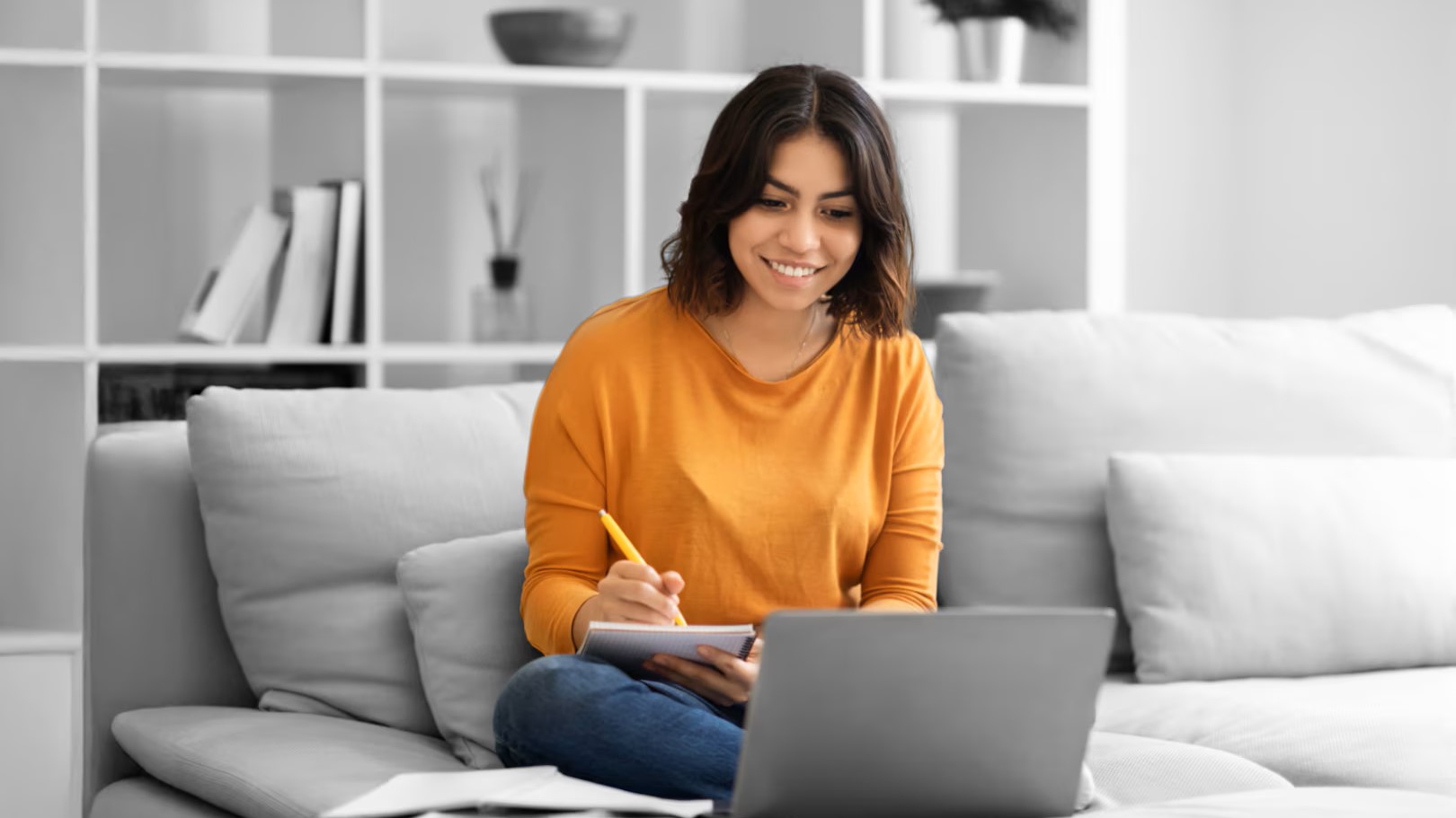 Woman sitting on sofa, smiling, looking at laptop and writing notes on a notepad. 