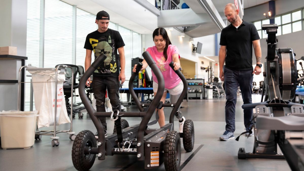 U.S. Air Force Senior Airman Hailey Alaniz, an aerospace medical service technician with the 2nd Medical Group, pushes a weighted sled alongside physical therapist Carey Wright as Staff Sgt. Donovan Lefever looks on during a functional rehabilitation session at the Center for the Intrepid at Brooke Army Medical Center, Joint Base San Antonio-Fort Sam Houston, Texas, Aug. 5, 2026. The all-surface wheeled sled uses progressive magnetic resistance that automatically increases with speed, allowing users to build lower-body power and cardiovascular endurance. Its bi-directional design provides a joint-friendly, low-impact training option ideal for physical therapy rehabilitation. (DoW photo by Jason W. Edwards)