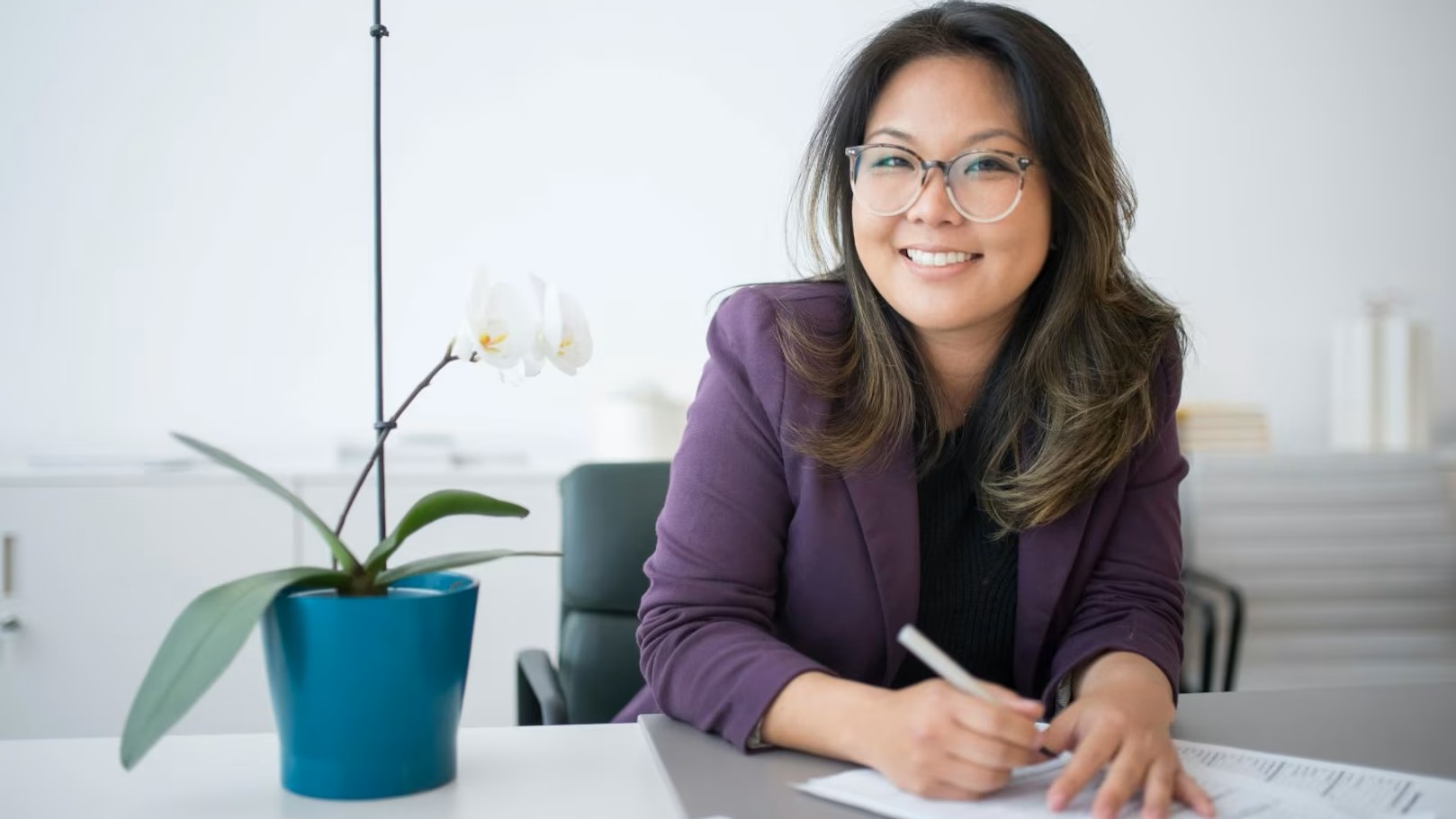 Smiling woman, sitting at a desk with an orchid plant next to her