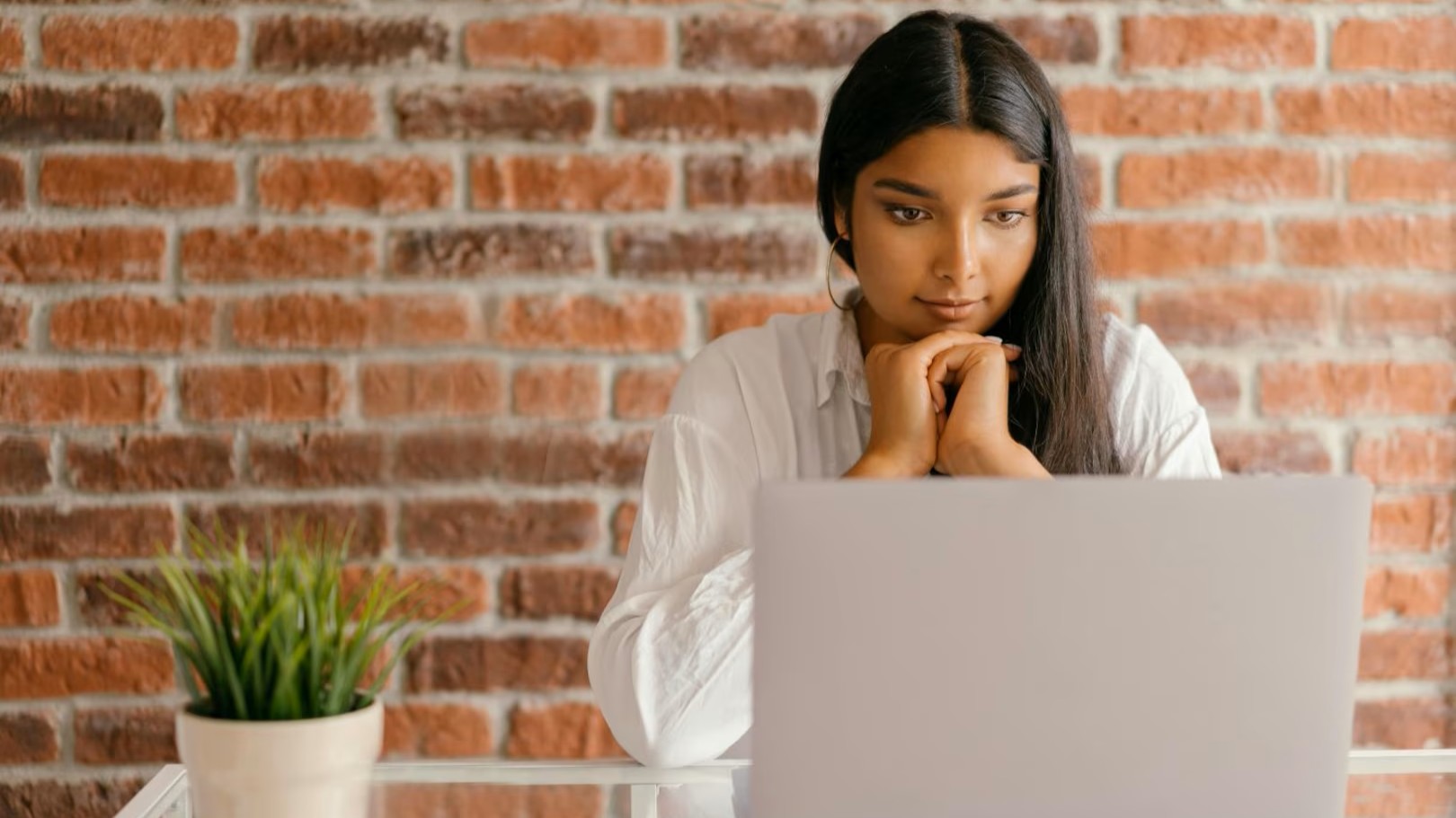 Woman sitting at desk looking at her laptop, with a brick wall behind her and a plant on the desk.