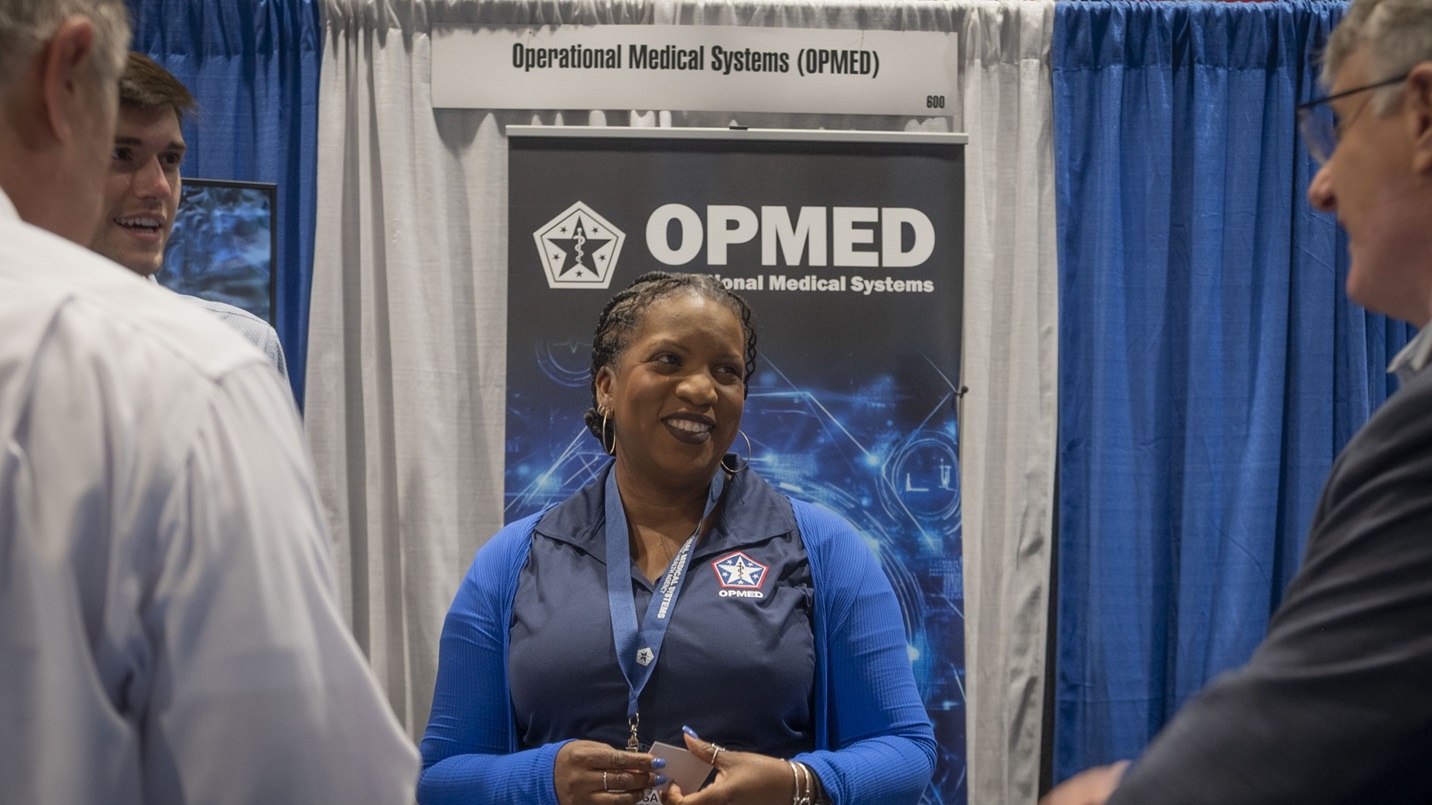 Jericsa Rogers, a logistics management specialist with the Operational Medical Systems Program Management Office, speaks with attendees of the Military Health System Research Symposium during exhibit hours, Kissimmee, Florida, Aug. 6, 2025.